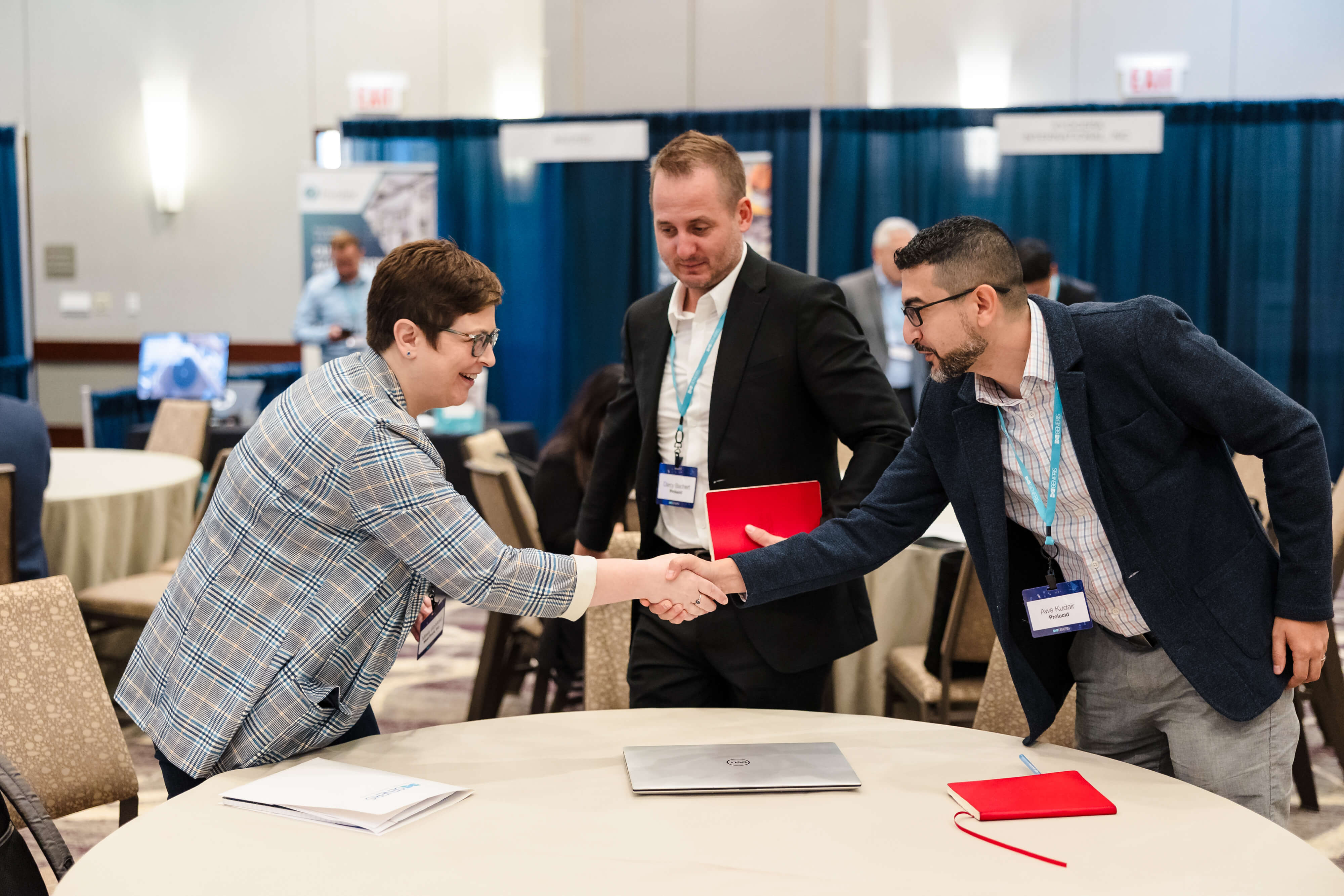 Three professionals at a conference room, actively engaged in a handshake. A woman in a checkered blazer with glasses is reaching across a table with a laptop to shake hands with a man in a navy jacket and glasses. Another man in a black suit holding a red folder observes their interaction. The background features conference booths and other attendees.