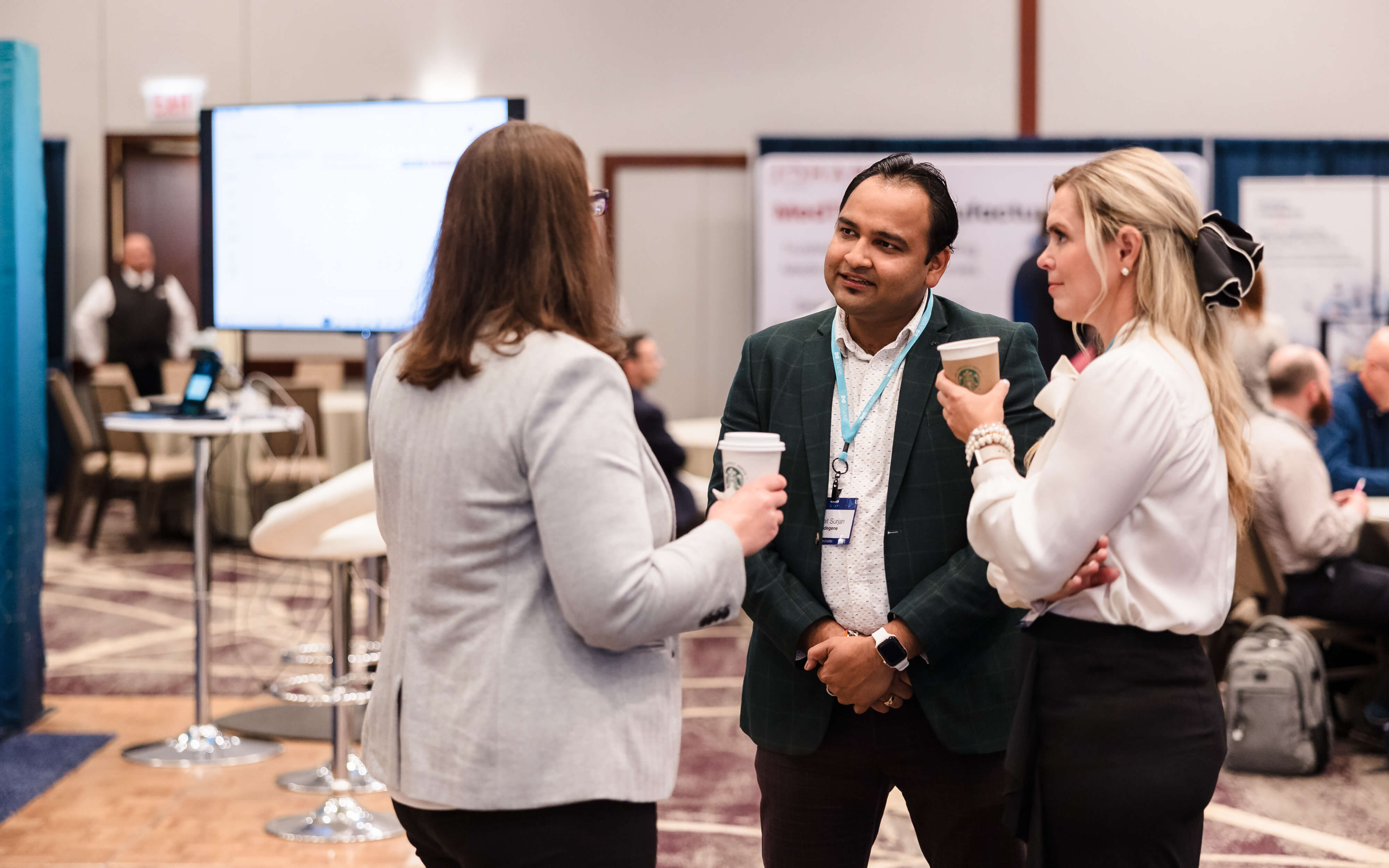 Three professionals in a conference setting, with two women and one man engaged in a conversation. The man in a green blazer holds a coffee cup and wears a name badge, while one woman, with her back to the camera, also holds a coffee cup. The other woman, with blonde hair, stands beside him, looking attentive. In the background, there are tables, chairs, and display screens, suggesting an ongoing event or presentation.