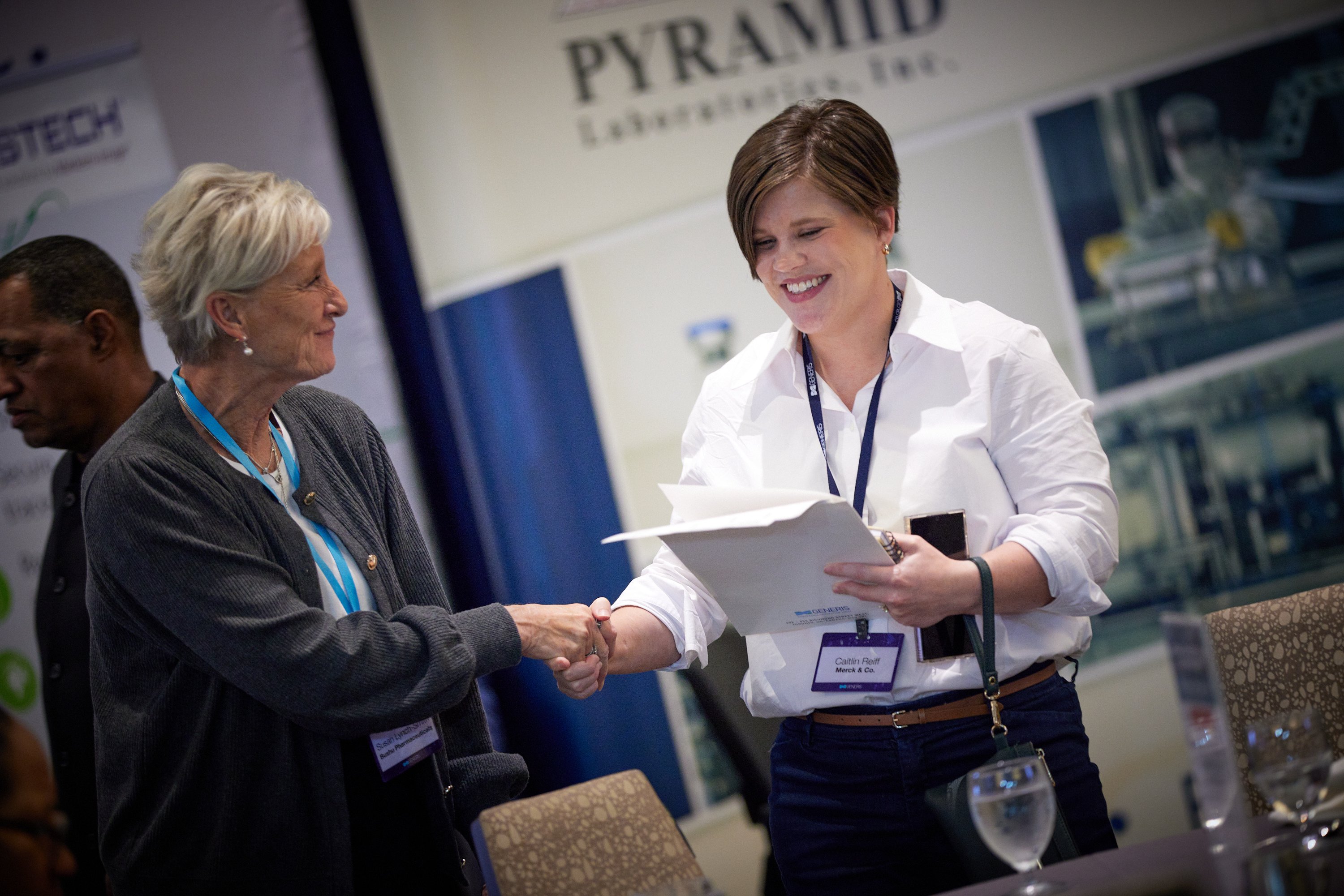 In the photo, two women are actively engaged in a conversation at a conference/ summit. An older woman with silver hair, wearing a gray cardigan, extends her hand to shake hands with a younger woman. The younger woman, with short brown hair, is wearing a white blouse and dark pants, holding a folder and has a name tag. In the background, there are banners of companies or sponsors, suggesting that this is a professional setting. A man is also partially visible on the left side of the image.